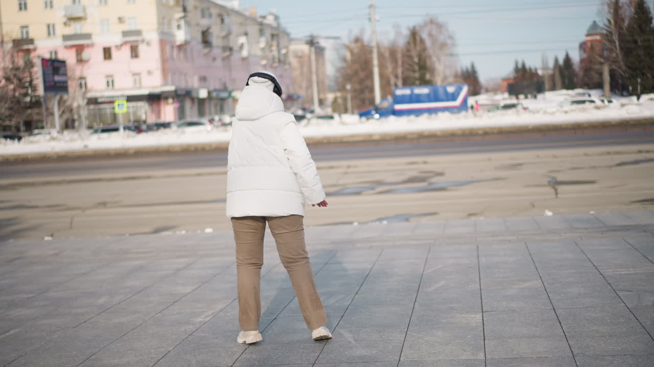 Back view of girl wearing white hooded winter jacket and headphones dancing joyfully on paved recreation ground in cold weather while cars drive by on road and pedestrians walk past