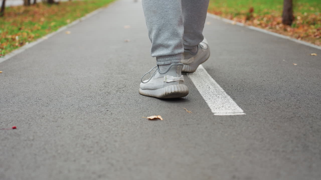 Leg view of person in sneakers and joggers walking briskly on tarred road with scattered autumn leaves as another person approaches in blurred distant background