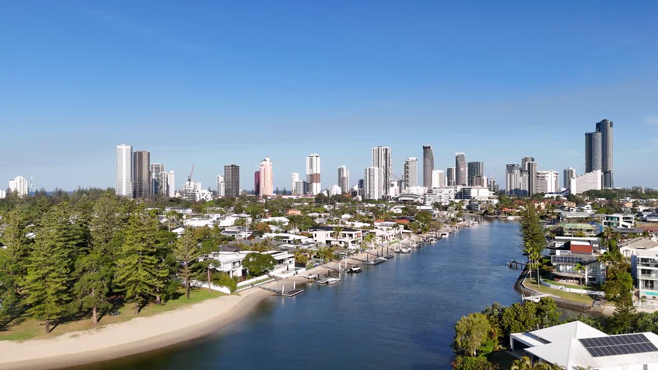 Drone glides over river, luxury homes, and city skyline under bright daylight, clear blue sky