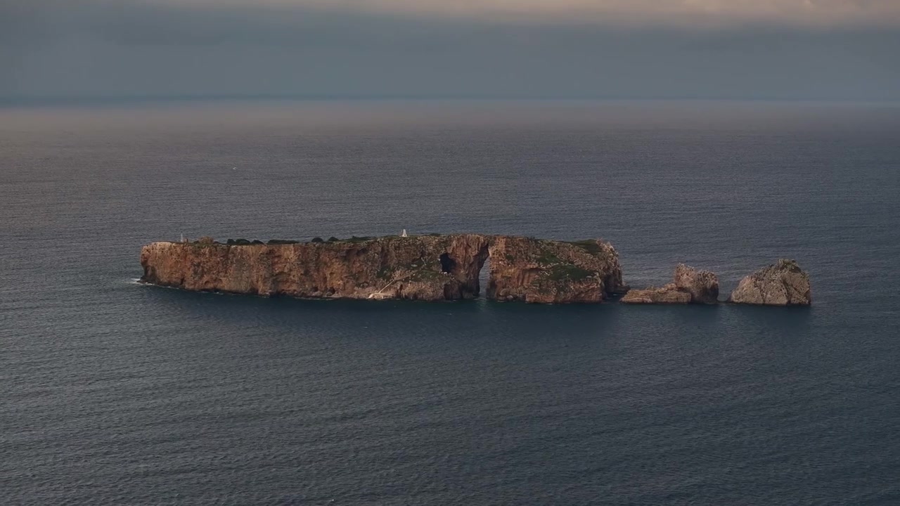 Luftfoto med teleobjektiv af klippeøen Tsichli Baba med dens berømte naturlige stenbue og franske mindesmærke, omgivet af dybblåt havvand under en stemningsfuld skumringshimmel nær Pylos, Grækenland.