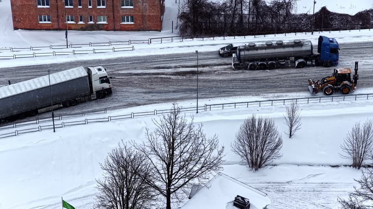 Aerial overhead view of heavy trucks and tanker stuck sliding on icy snowy road during heavy snowfall in Latvia with snowplow working nearby residential buildings winter conditions