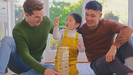 family with two dads playing game with daughter at home stacking wooden bricks into tower