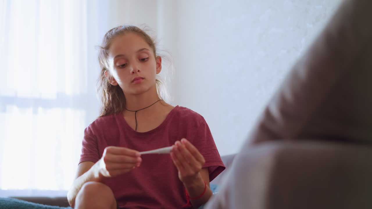 Little girl wearing maroon shirt and black choker carefully checks thermometer reading while attending to sick person at home showing concern focus and responsibility