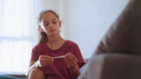 little girl wearing maroon shirt and black choker carefully checks thermometer reading while attending to sick person at home showing concern focus and responsibility