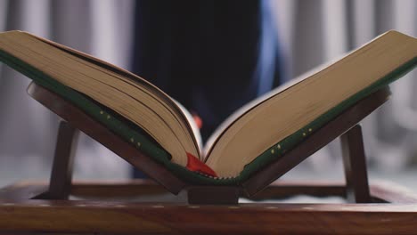 close up of open copy of the quran on stand at home with woman sitting behind