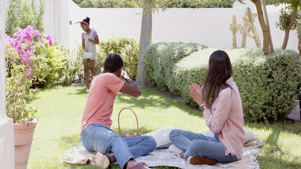 Easter, diverse family enjoying picnic on grass while child playing, relaxing in garden