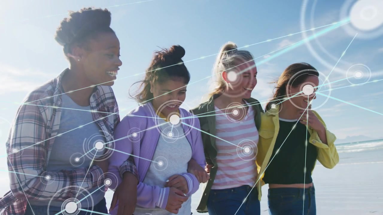 Four women linked arms walking beach, nodes growing, tilting camera, lifting heads, showing tech