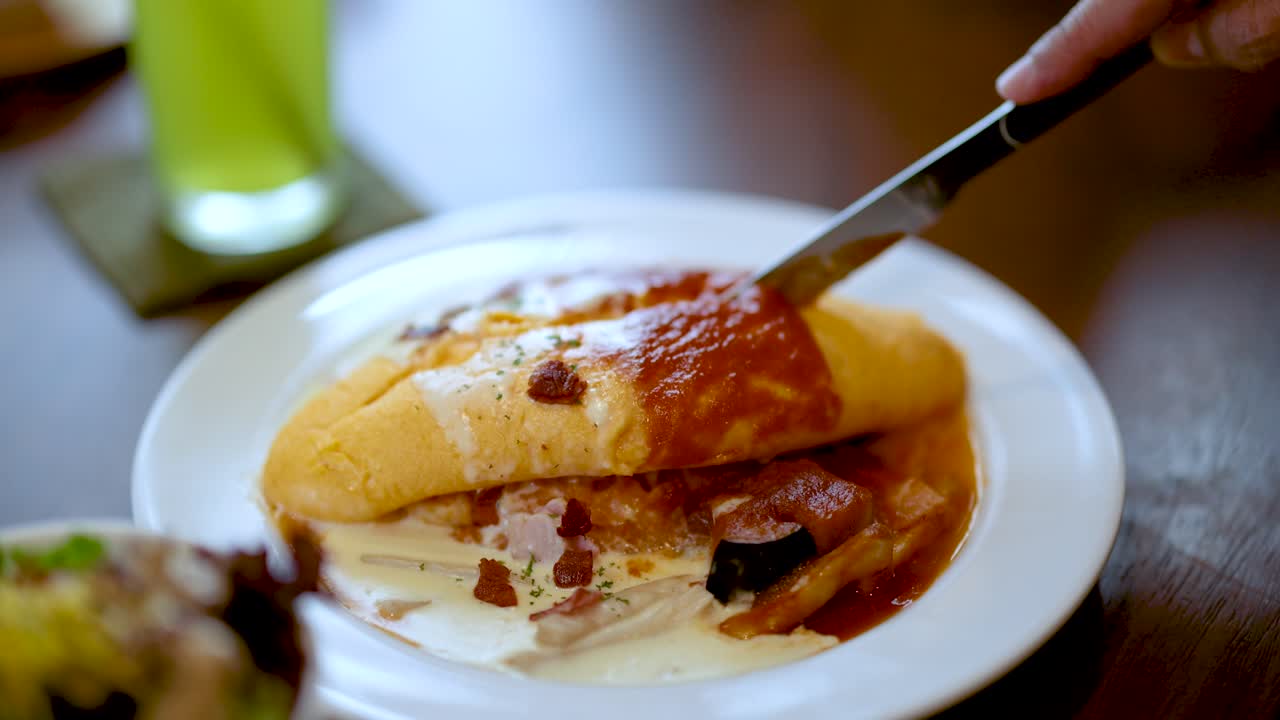 Hand slices omurice with knife on plate, natural daylight, shallow focus, casual dining setting