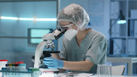trabajadora de laboratorio en uniforme de protección mirando a través de un microscopio
