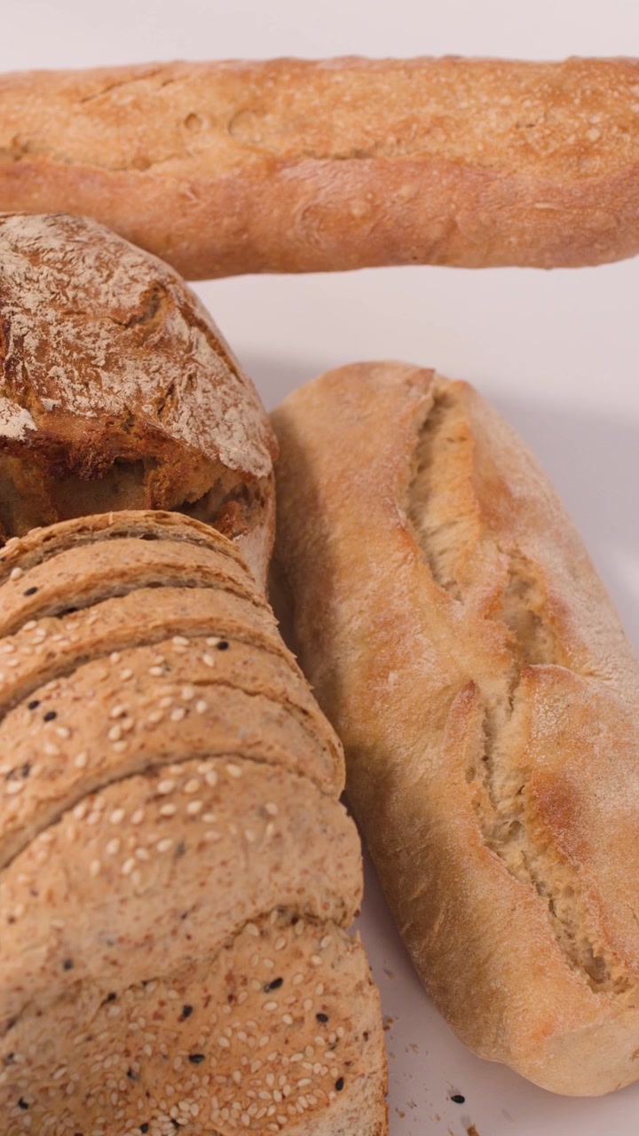 Lenta panoramica della telecamera su una varietà di pane a lievitazione naturale, baguette e cereali a fette su una superficie bianca