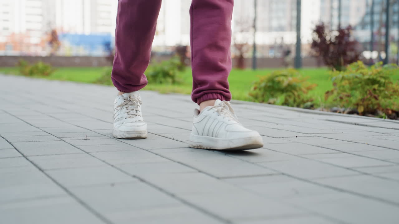 Close up leg view of walking woman in her twenties wearing white sneakers and burgundy jogger trousers moving along gray tiled pavement in urban park setting capturing dynamic stride detail
