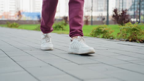close up leg view of walking woman in her twenties wearing white sneakers and burgundy jogger trousers moving along gray tiled pavement in urban park setting capturing dynamic stride detail