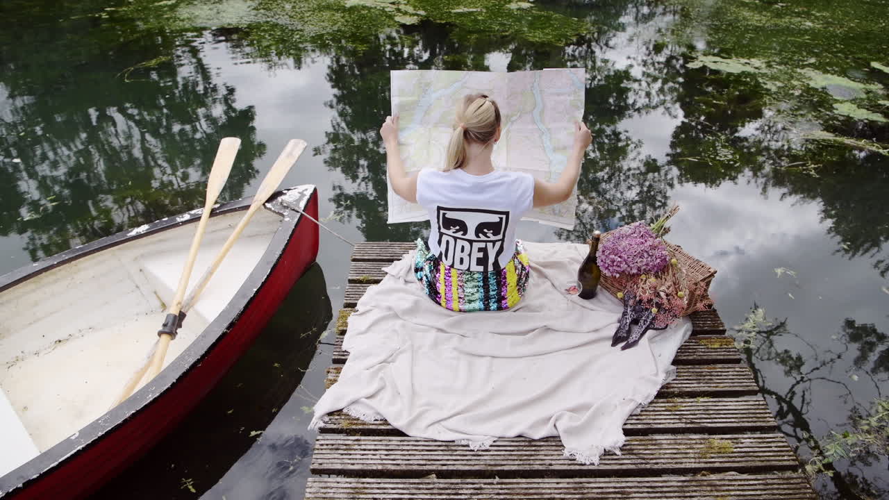 Woman holding large map while sitting on wooden jetty, relaxing by the water, picnic for one, oars and a traditional rowboat at lake, static shot