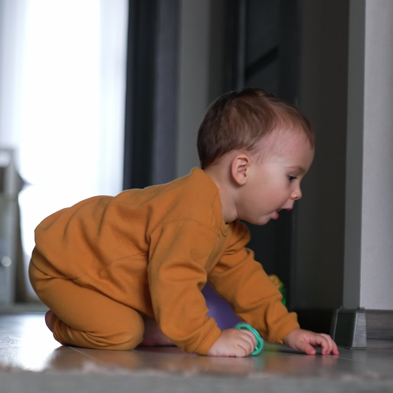 Nice little kid sits on the floor with his toys beside. Baby boy starts to crawl and yawns approaching camera. Blurred backdrop