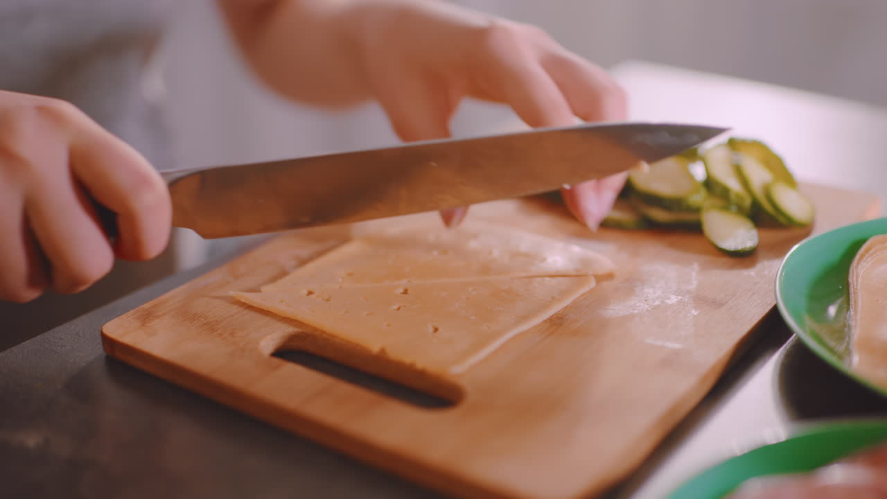 Close up of lady holding cheese slice on wooden cutting board with fresh cucumber slices beside plate, preparing ingredients for sandwich in bright kitchen atmosphere with attention to detail