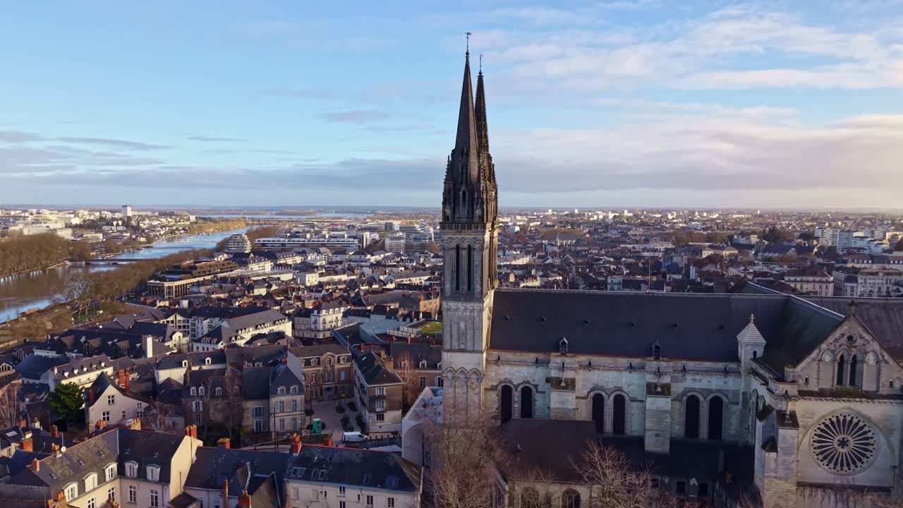 La Cathédrale Saint Maurice d’Angers nei Paesi della Loira, con vista sul fiume Maine e sul denso tessuto urbano di Angers, mostra l'architettura gotica angioina e le sue doppie guglie contro un cielo limpido, drone in ascesa.