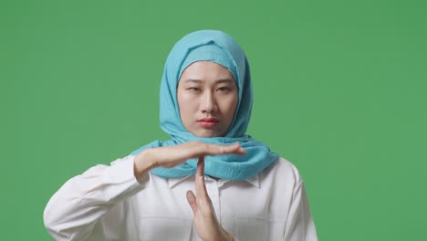 close up of asian muslim woman looking at camera and showing time out gesture while standing in the green screen background studio