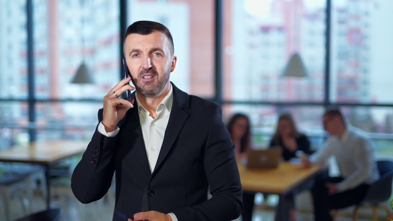 Man speaking emotionally on the phone and gesturing to the camera. Unpleasant business talk. Employees in blur at backdrop.