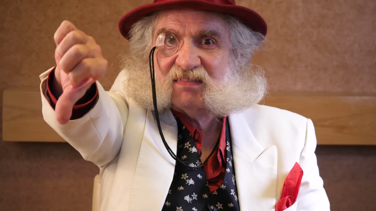 턱수염을 기른 노인이 단안경을 착용하고 카메라를 향해 엄지손가락을 아래로 내리며