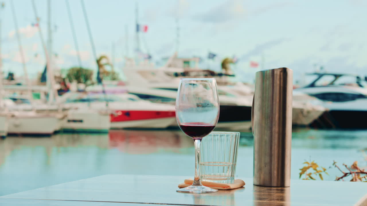 A half full glass of red wine stands on a restaurant table overlooking a calm marina with luxury yachts in soft focus
