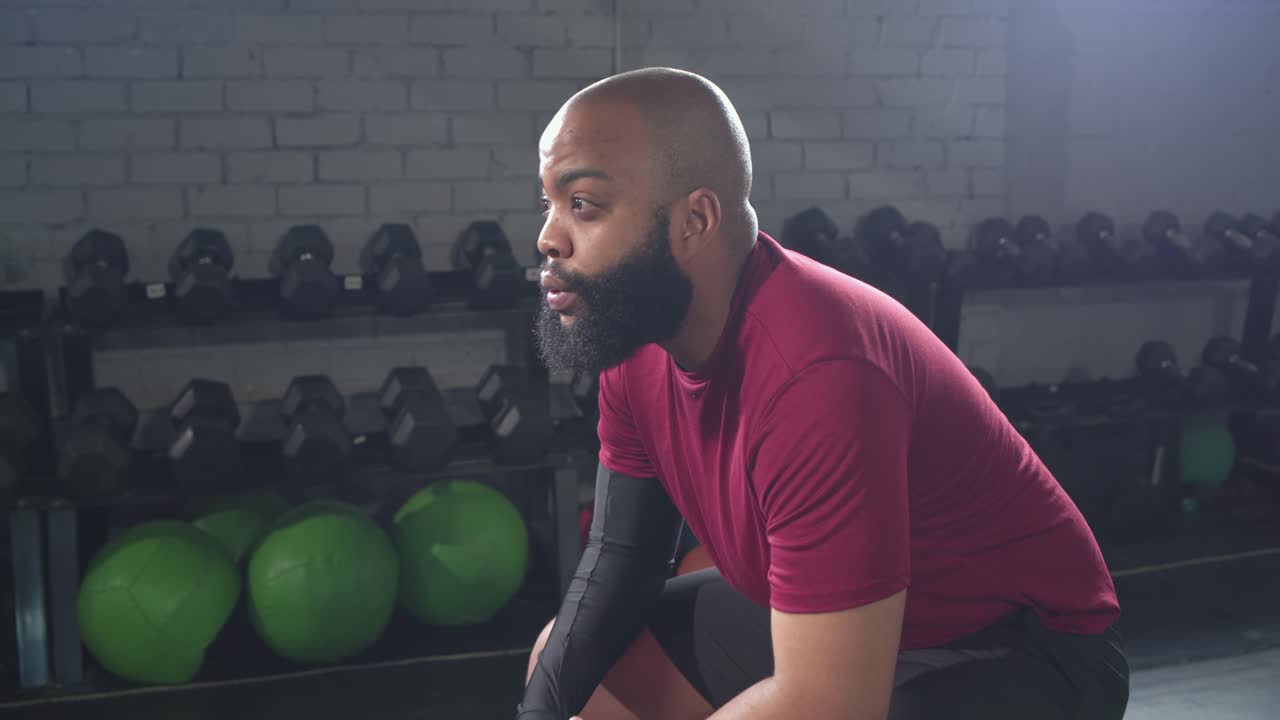 African American man in red shirt resting between sets on bench in gym, looking toward camera