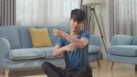 young asian athletic male in sports clothes sitting on a yoga mat and doing shoulder cross-arm stretch at home