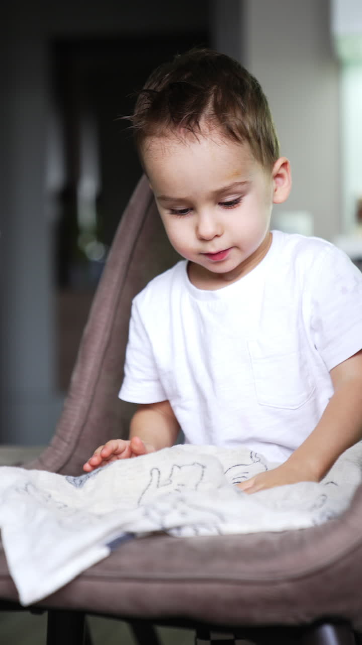 Little boy takes a towel and walks by the kitchen. Kid puts the towel on the chair and smiles to camera. Vertical video.