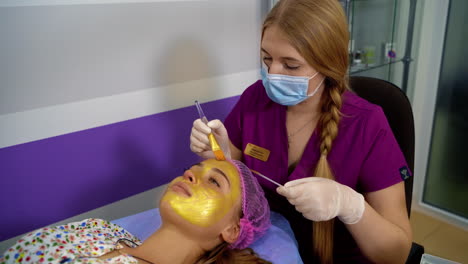 cosmetologist apply gold mask with brush on young pretty woman`s face in beauty clinic. spa salon. beautician making beauty facial skincare procedure.