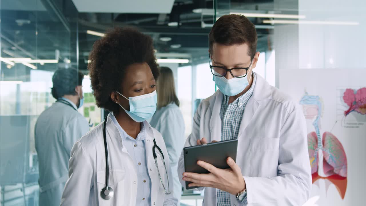 African american and caucasian male and female doctors in medical masks standing in hospital office discussing health problem and typing on tablet