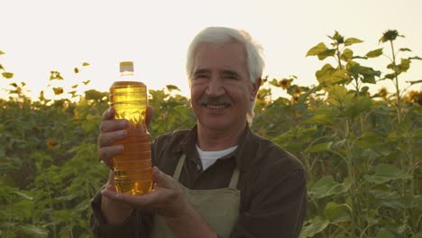 Zeitgenössischer-älterer-Grauhaariger-Mann-In-Schürze-Und-Hemd,-Der-Eine-Flasche-Sonnenblumenöl-Betrachtet,-Während-Er-Auf-Dem-Feld-Steht