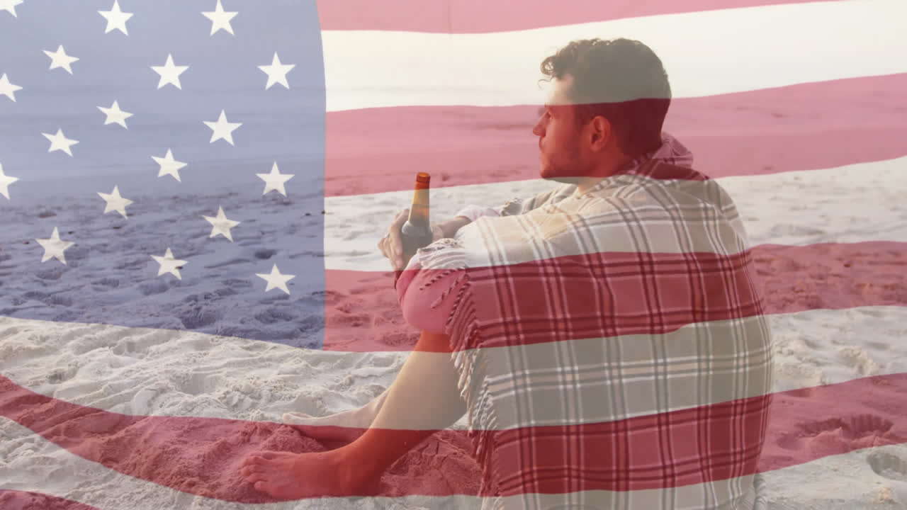 Animation of american flag waving over man wrapped in blanket drinking beer on beach