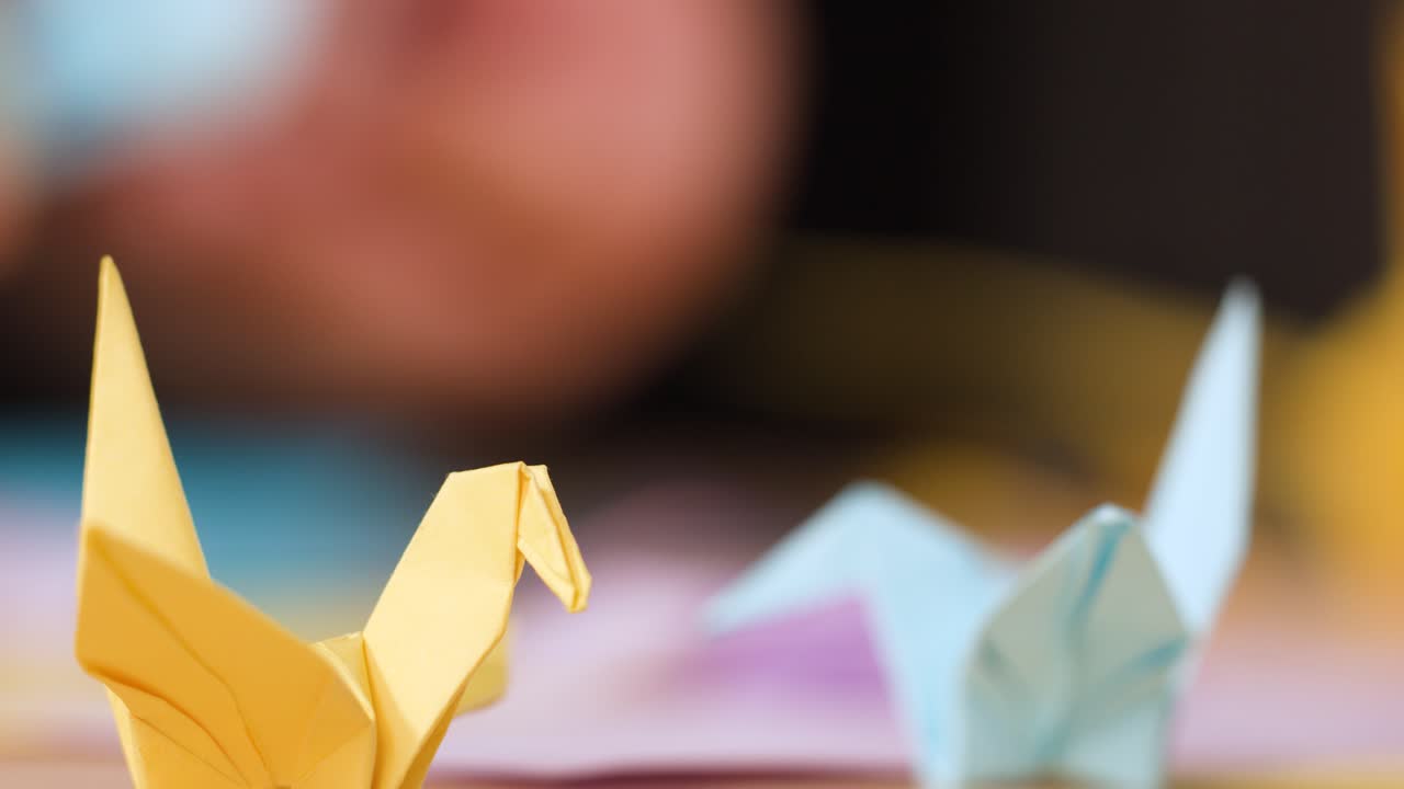Una gru di carta gialla è a fuoco, mentre delle mani piegano origami sullo sfondo sfocato