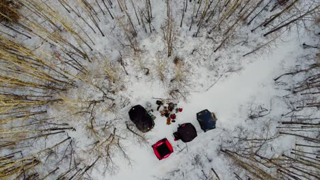 aerial birds eye view rise over father and son winter camping with summer and ice fishing tents forming a v with a manmade campfire dogs entertained on a sunset with bare trees tipped in yellow 2-3