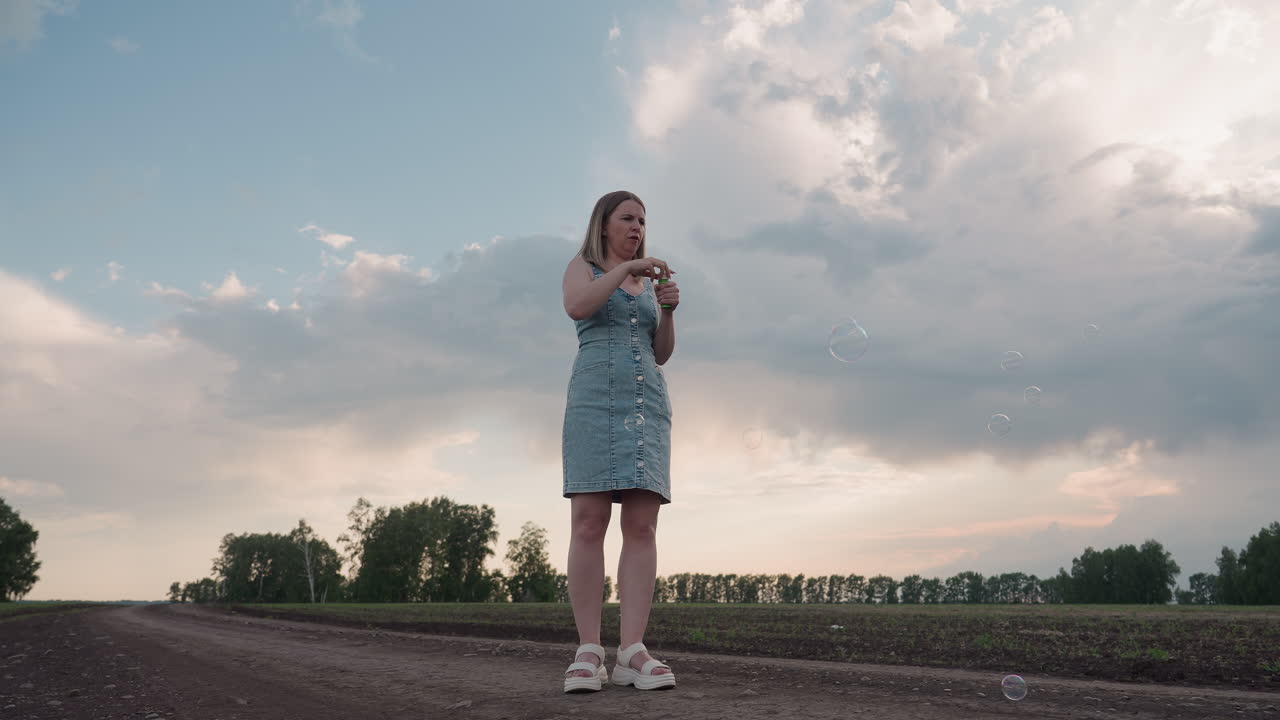 low angle view of young woman standing in open farmland blowing multiple iridescent soap bubbles into cloudy sky under soft evening light