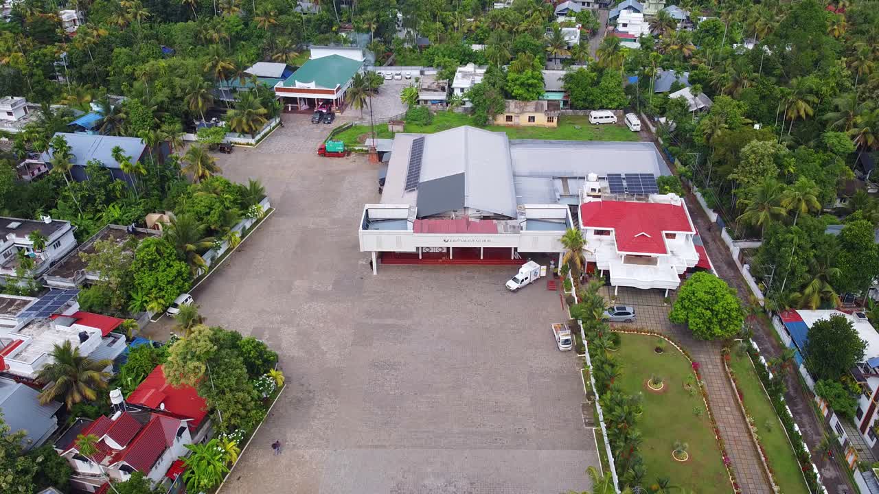Aerial view of a large event center or marriage hall with a spacious paved lot, surrounded by lush green trees and tropical residential buildings