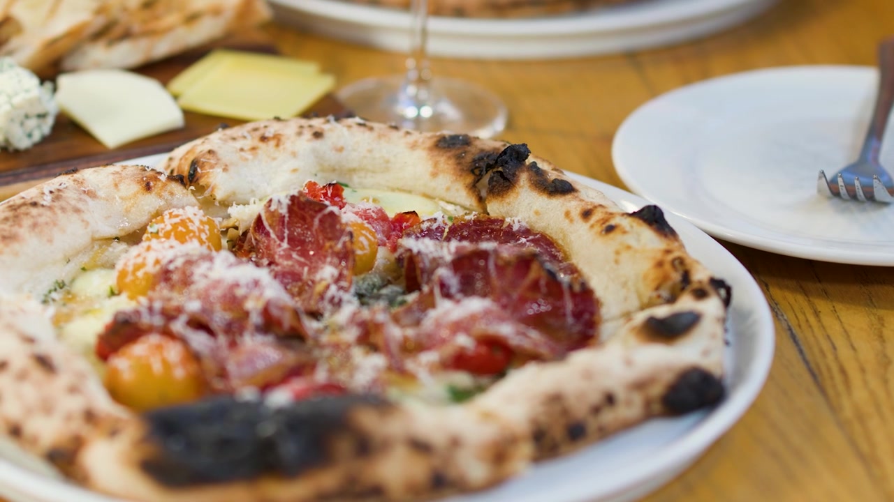 Close-up de mãos usando uma faca para cortar pizza fresca de presunto cru em uma mesa de madeira