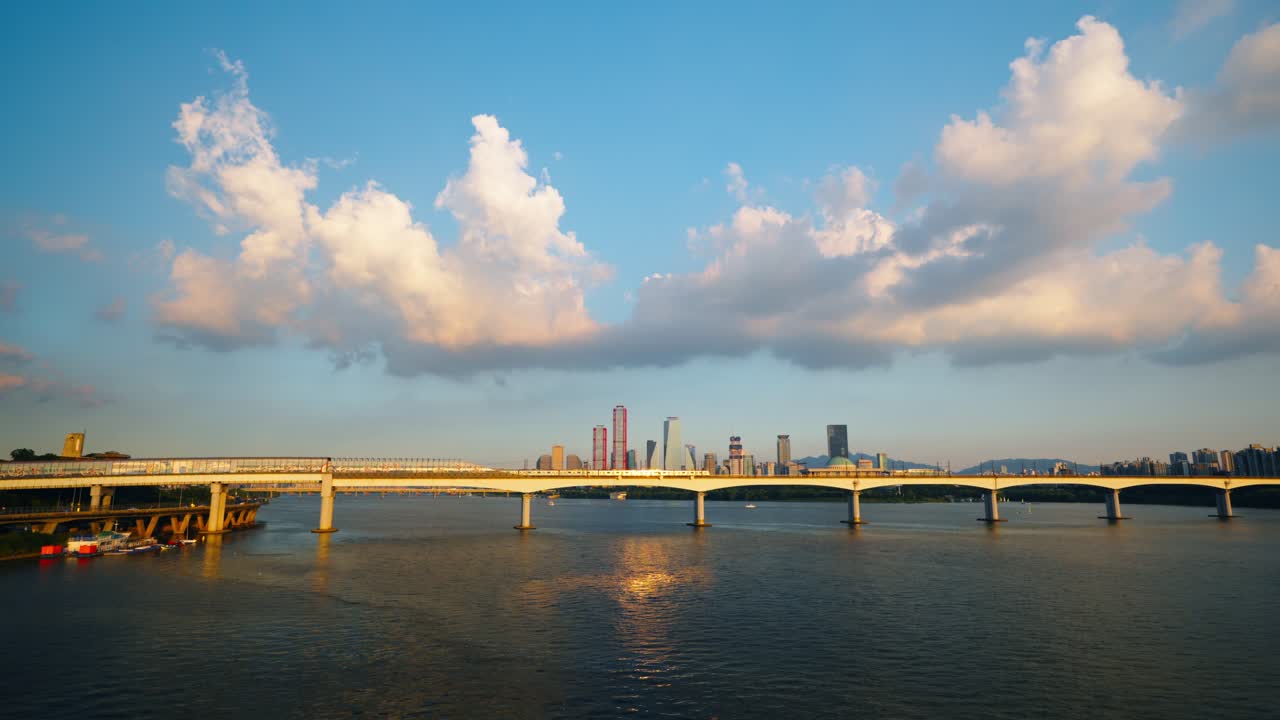 A stunning aerial view shows a metro train crossing the Dangsan Railway Bridge over the Han River in Seoul, with the modern skyscrapers of the Yeouido business district in the background at sunset