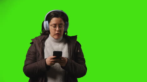 young woman wearing wireless headphones streaming music from mobile phone walking into frame against studio green screen