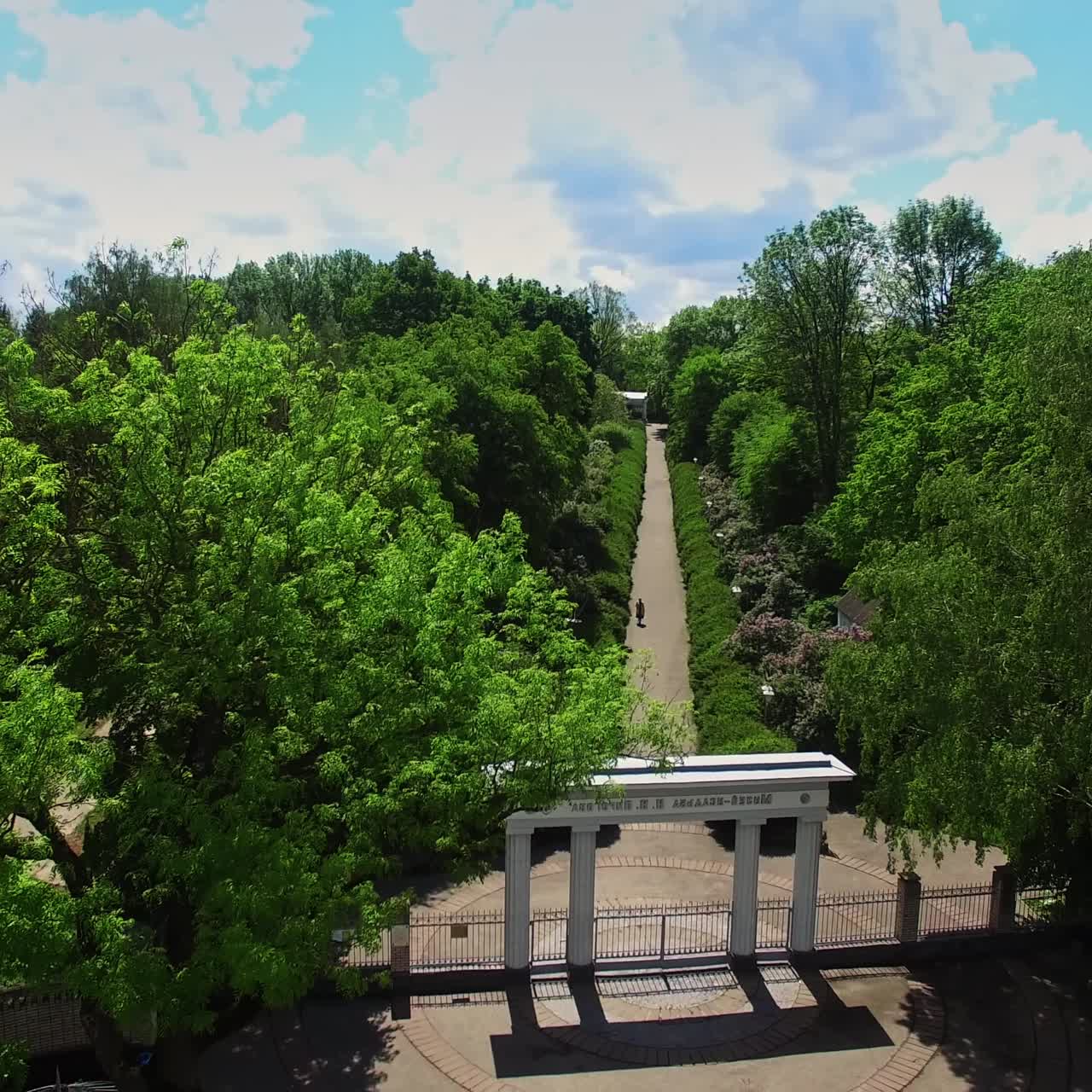 White columns at the entrance to the beautiful green park. Drone footage over the car road and lawn. Sunny day backdrop