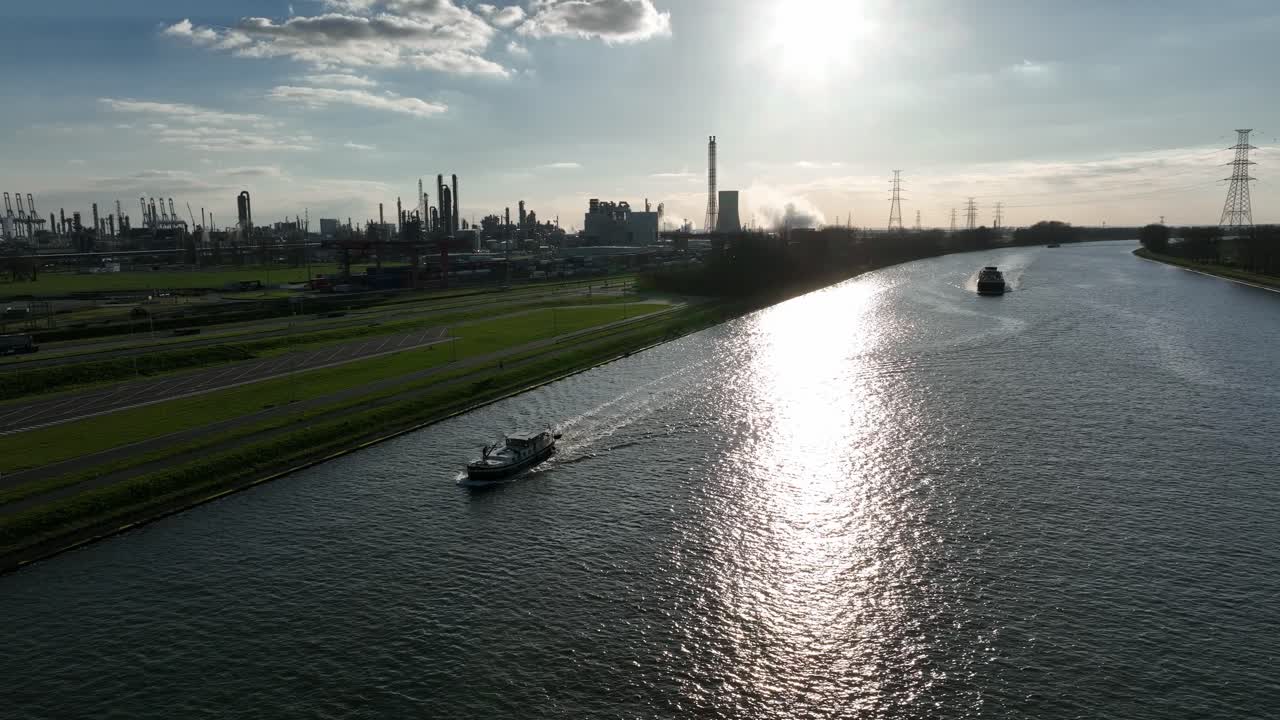 Zoom in aerial shot of cargo boat sailing Antwerp industrial canal with factories, chimneys, and pipelines during sunset light