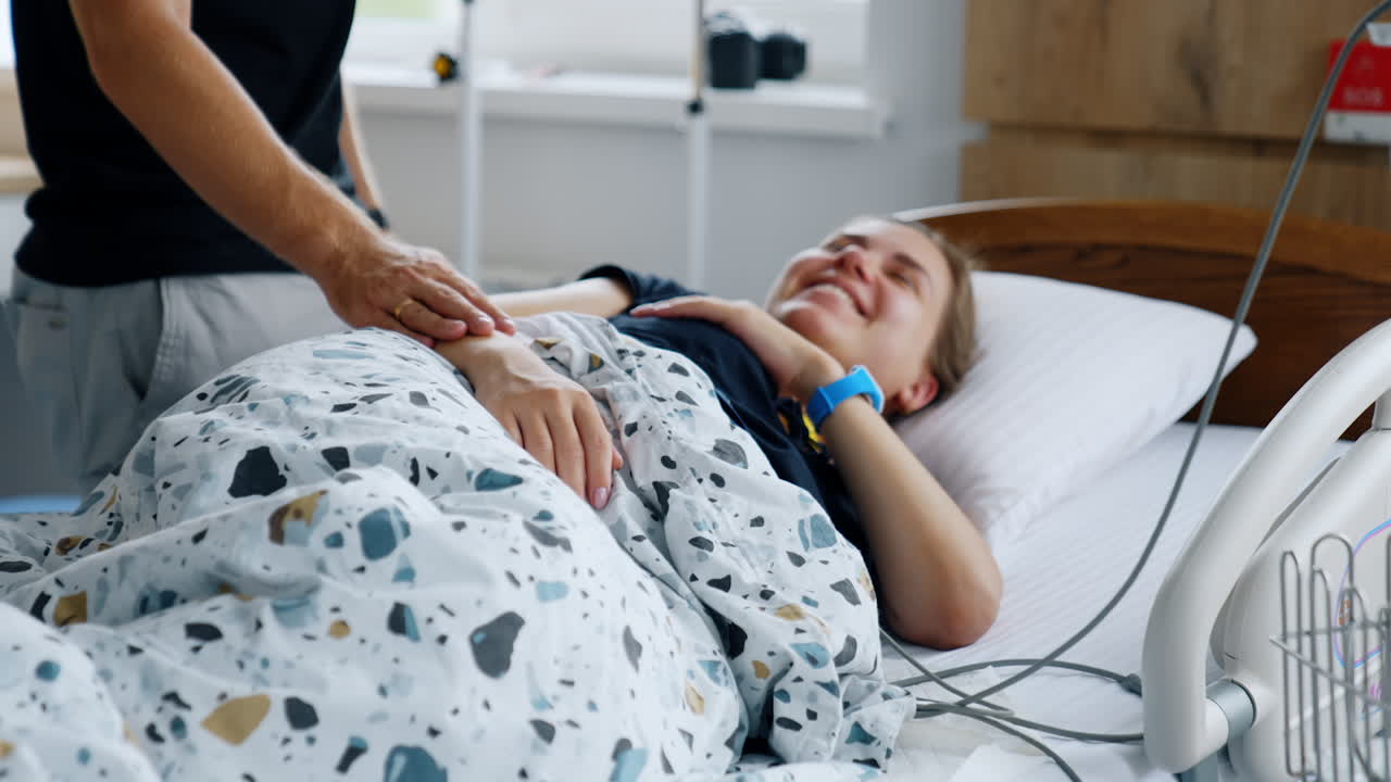 Caucasian woman expecting a child lies in hospital ward. Man standing beside stroking her arm.