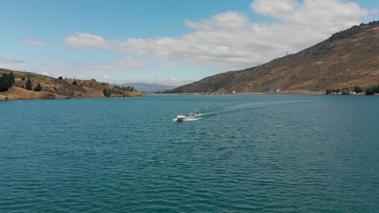 cámara lenta: esquí acuático detrás de un bote en el lago dunstan cerca de la represa clyde, otago central, nueva zelanda con montañas y nubes en el fondo