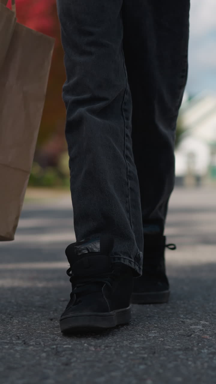 primer plano de piernas caminando en un área residencial, persona cargando una bolsa de papel, con árboles de otoño, casas y un coche estacionado en el fondo, capturando un día de otoño suburbano
