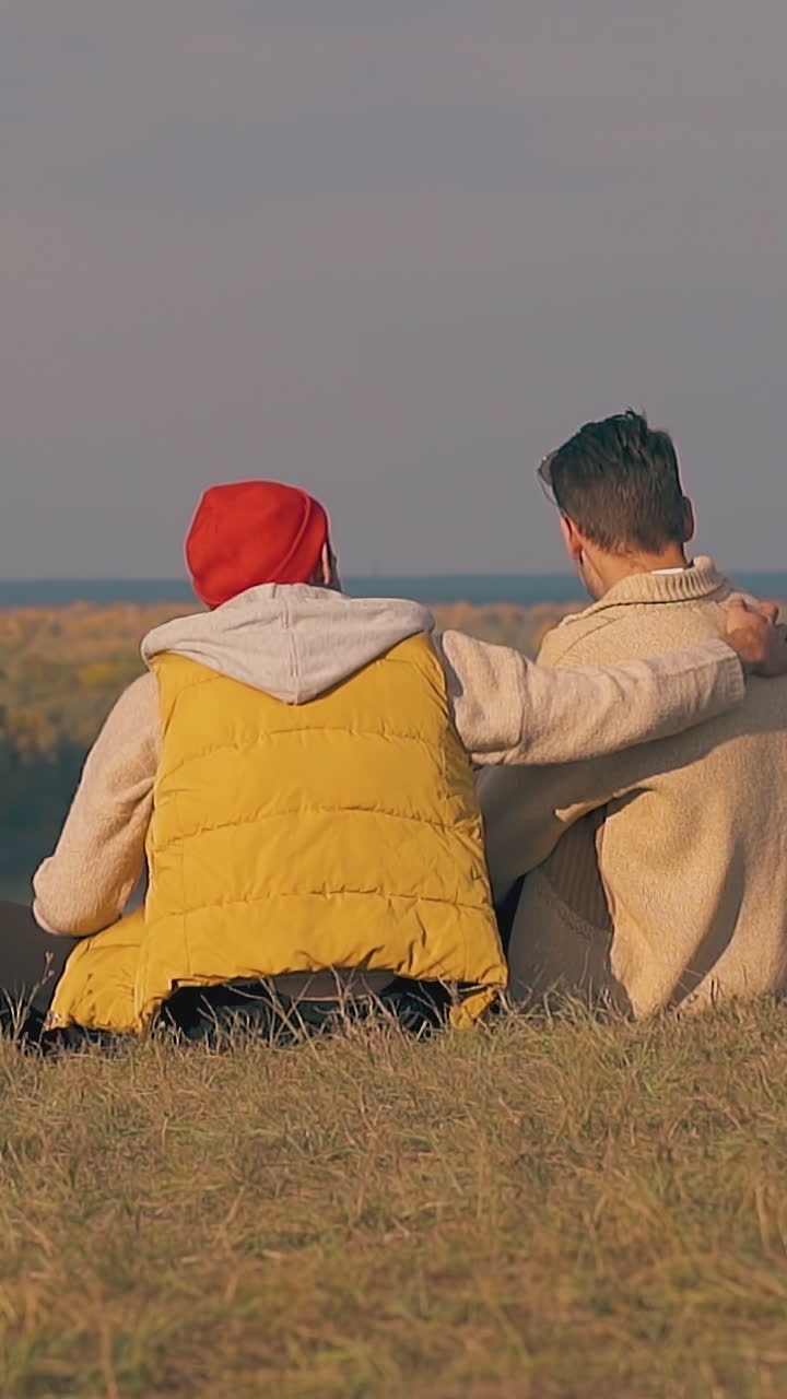 man sits near boyfriend and hugs on river bank grass edge in early morning slow motion backside view