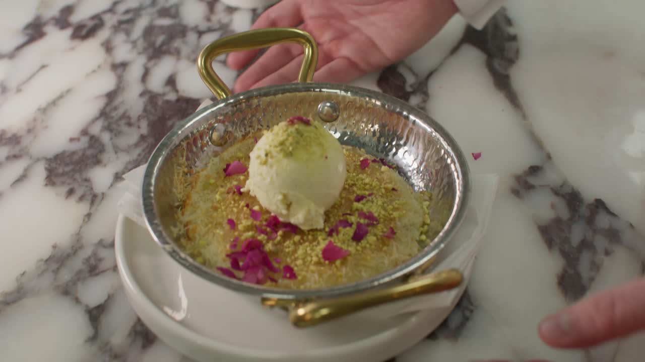 A cinematic close-up shot of a chef's hand using an ice cream scoop to place a perfect dollop of vanilla ice cream onto a warm Kunafa dessert, beautifully garnished with crushed pistachios and dried r
