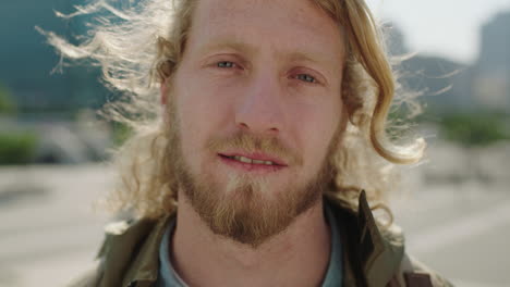 close up portrait of young blonde bearded man pensive student looking calm at camera urban city background