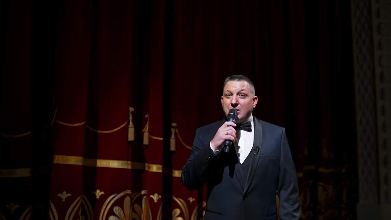 Speaker on stage. Male host in elegant costume speaking into a microphone looking on camera on the background of red curtains in the theater. Slow motion.