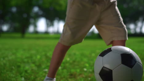 closeup kid feet kicking up football ball. boy play on green lawn in park.