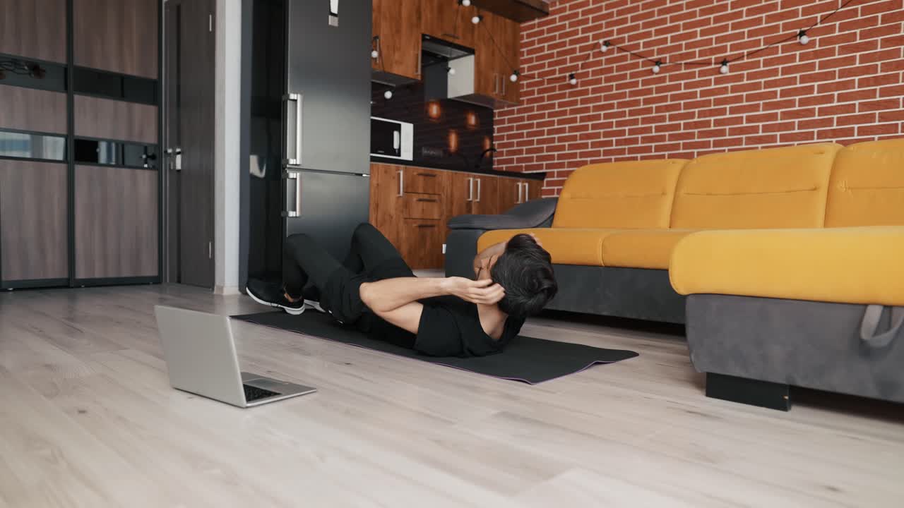 Man training on mat with laptop at home, doing bicycle exercise on back