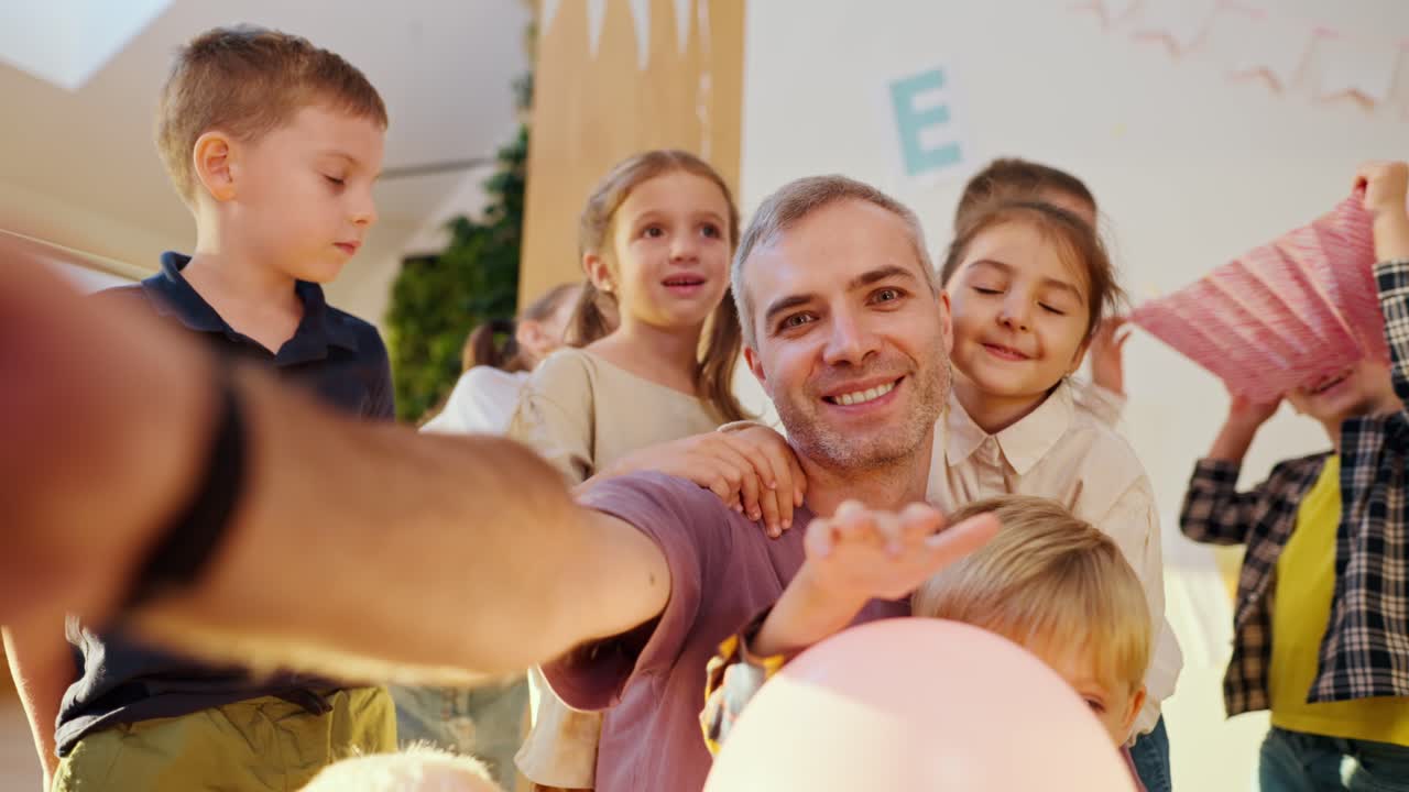 보라색 티셔츠를 입은 회색 머리카락을 가진 행복한 남성 교사는 학교를 준비하는 클럽에서 학생들과 셀피를 찍습니다.